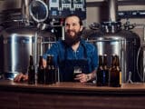 Um homem sorridente, com barba grande, usando camisa azul e avental preto, com várias garrafas de cerveja no balcão a sua frente