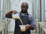 Homem em uma cervejaria artesanal despeja líquido âmbar entre recipientes transparentes, ao lado de tanques de fermentação em inox, ilustrando o processo que envolve a levedura da cerveja na produção