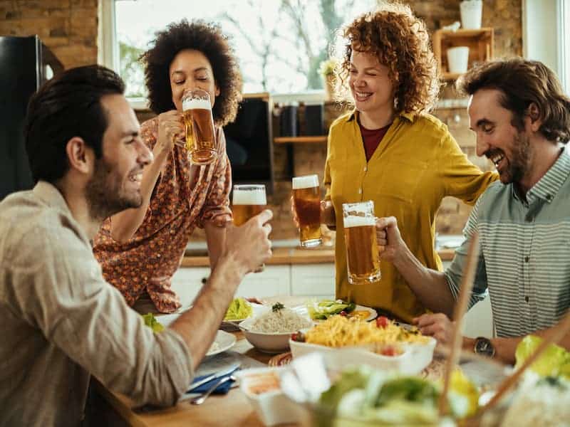 A imagem mostra um grupo de quatro amigos reunidos ao redor de uma mesa, sorrindo, conversando e brindando com copos, pensando em beber ou degustar cerveja