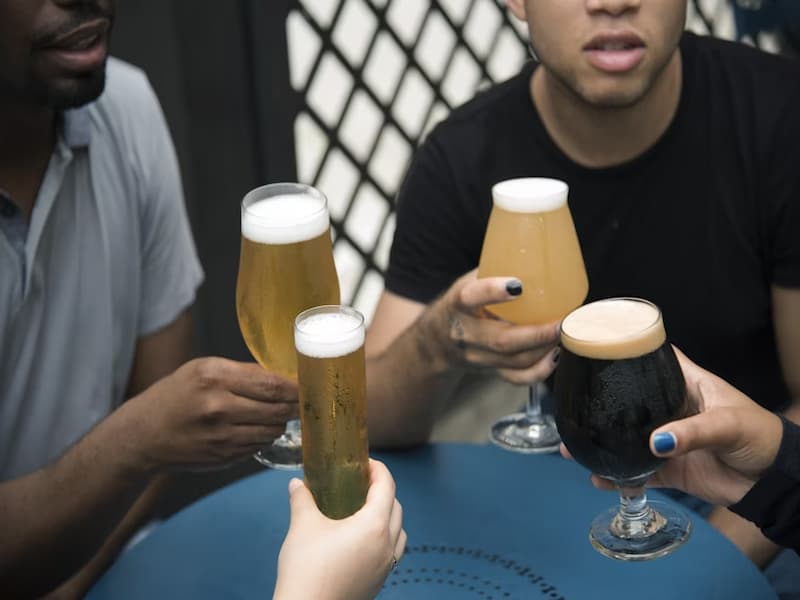 A imagem mostra um grupo de pessoas brindando com diferentes tipos de cerveja artesanal, sentadas ao redor de uma mesa azul, vendo se vão beber ou degustar cerveja