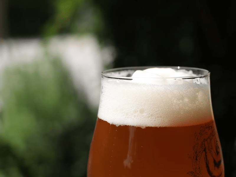 A imagem mostra um copo de cerveja clara (possivelmente uma IPA ou uma Belgian Ale) servido com uma camada de espuma branca no topo — o chamado colarinho. A espuma é fina, com bolhas pequenas e bem distribuídas, formando uma cobertura uniforme.