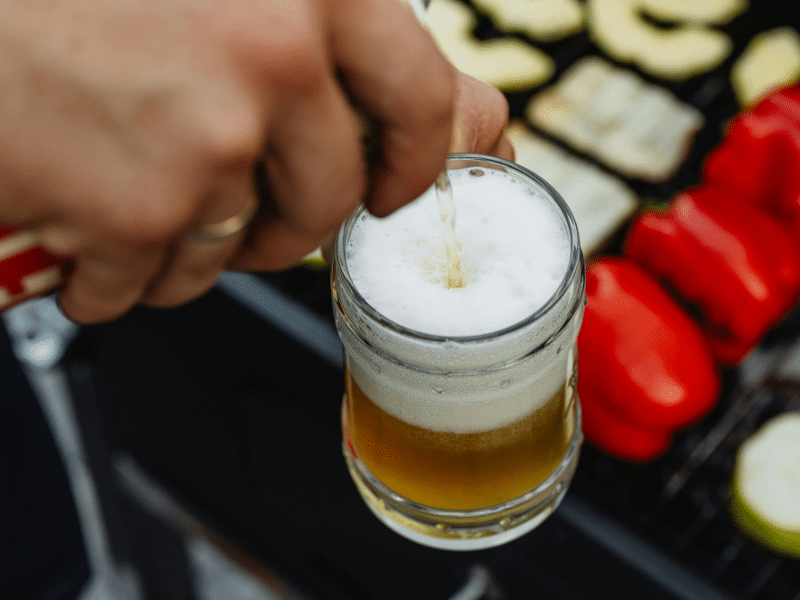 A imagem mostra uma cena típica de um churrasco ao ar livre. Em destaque, uma mão está segurando uma caneca de vidro transparente enquanto a cerveja está sendo servida, criando uma camada de espuma branca (colarinho) na parte superior do copo. A cerveja tem uma coloração clara e dourada.