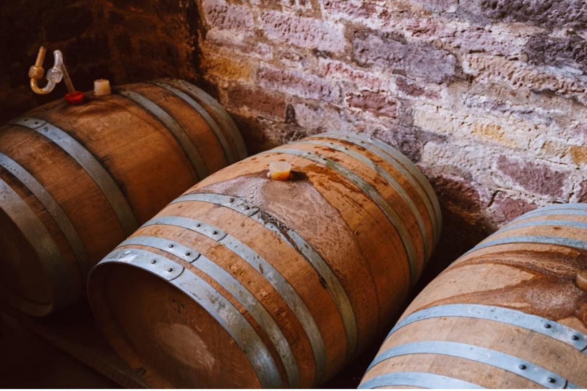 A imagem mostra um ambiente rústico e tradicional de maturação de bebidas alcoólicas em barris de madeira, provavelmente em uma adega subterrânea ou porão de envelhecimento.