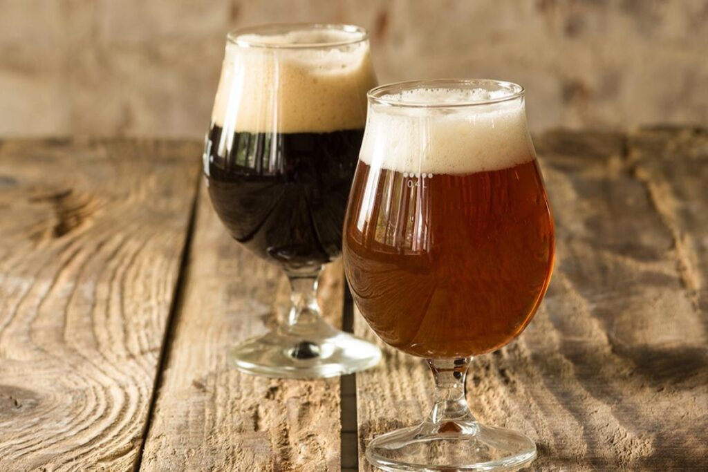 A imagem apresenta dois copos de cerveja artesanal servidos em copos do tipo tulipa, dispostos sobre uma mesa de madeira rústica. O cenário é simples e aconchegante, evocando uma atmosfera de apreciação e degustação.