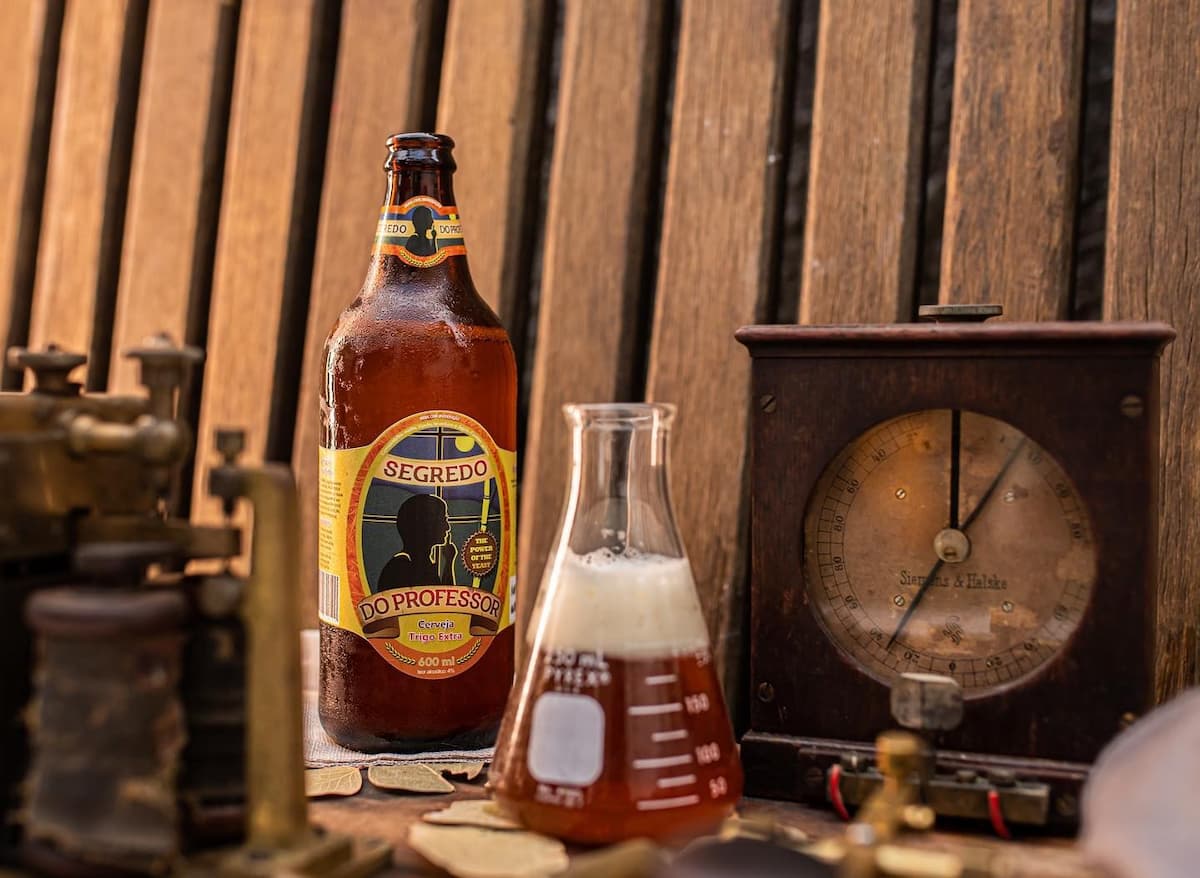 A imagem retrata uma das cervejas artesanais nas cidades cervejeiras no Brasil. Em destaque, uma garrafa da cerveja “Segredo do Professor”, com rótulo criativo e chamativo que evoca um ar enigmático e intelectual — reforçado pela silhueta de um personagem com óculos e dedo sobre os lábios, como quem guarda um segredo.
