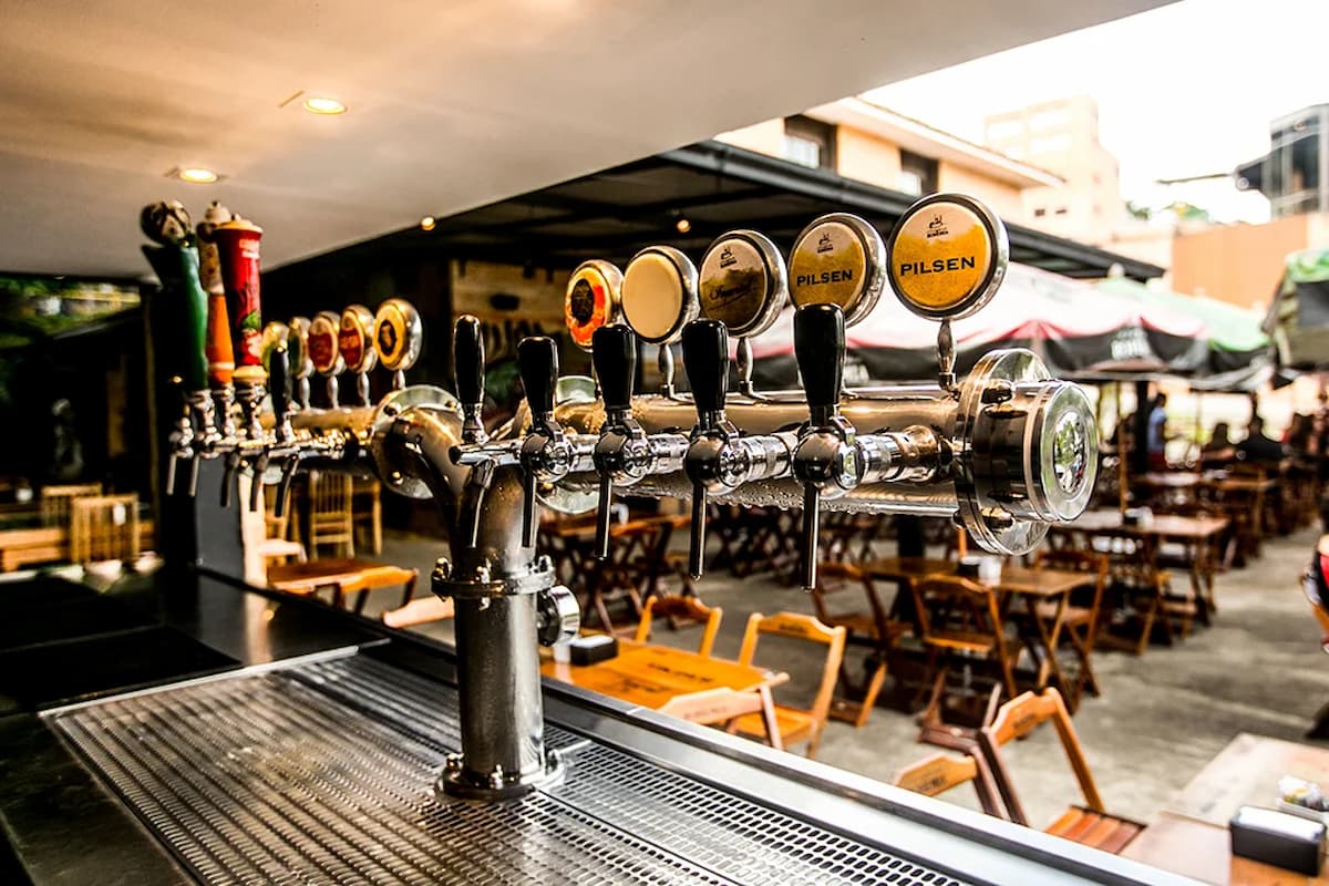 A imagem mostra uma fileira de torneiras de chope instaladas em um bar ao ar livre, destacando um ambiente típico de cervejarias artesanais brasileiras. As torneiras, com rótulos variados — incluindo o estilo "Pilsen" —, sugerem a oferta de diferentes tipos de cervejas, o que é característico do crescente mercado nacional de cervejas especiais e artesanais.