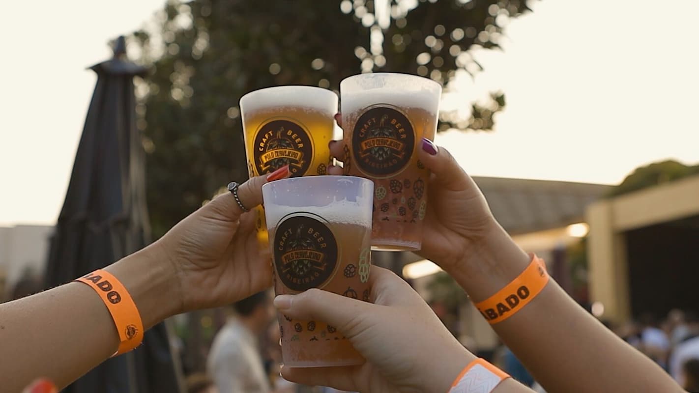 A imagem mostra quatro pessoas brindando com copos de cerveja artesanal durante um evento ao ar livre, no que parece ser um festival de cervejas.