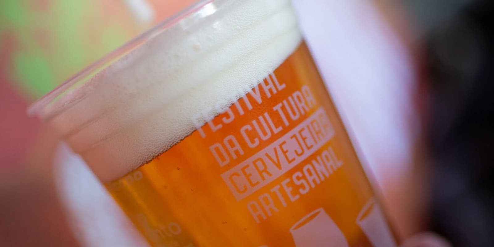 A imagem mostra um copo de plástico translúcido cheio de cerveja artesanal, com uma coloração dourada e uma generosa camada de espuma cremosa no topo. No copo, está estampado o texto "Festival da Cultura Cervejeira Artesanal", reforçando o contexto de um evento dedicado à celebração da produção local e independente de cervejas no Brasil.