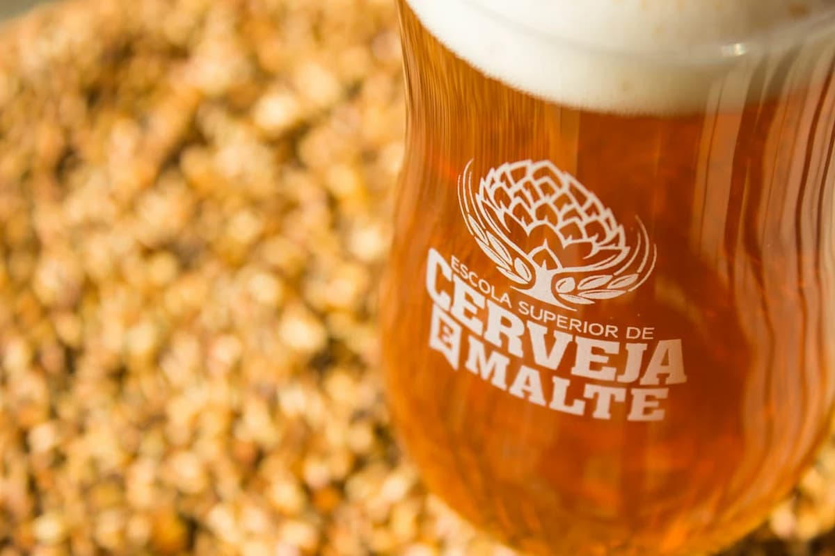 A imagem mostra um copo de cerveja artesanal com espuma cremosa e líquido âmbar, estampado com o logotipo da Escola Superior de Cerveja e Malte (ESCM), uma das principais instituições de ensino nas cidades cervejeiras no Brasil