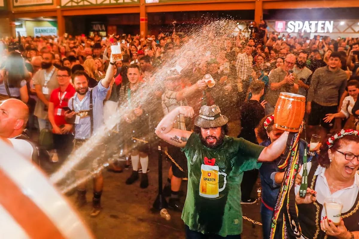 A imagem retrata uma grande celebração popular nas cidades cervejeiras no Brasil, claramente inspirada na cultura germânica. Um grande número de pessoas está reunido em um ambiente festivo, muitas vestindo trajes típicos alemães, como suspensórios, chapéus bávaros e vestidos tradicionais.