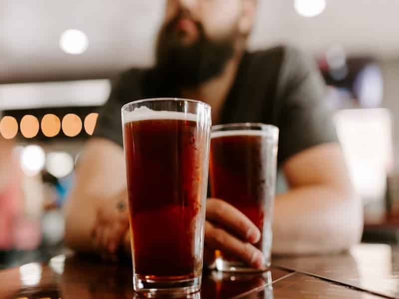 A imagem mostra um homem com barba sentado em um bar, segurando um copo de cerveja escura sobre a mesa. Outro copo semelhante está à frente dele. O foco está nos copos de cerveja — provavelmente uma Amber Ale, Porter ou Red Ale — que possuem coloração avermelhada a marrom, com pouca formação de espuma.