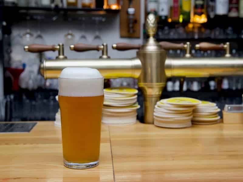 A imagem mostra um copo de cerveja artesanal servido sobre um balcão de madeira em um ambiente de bar ou pub. A cerveja tem uma coloração dourada turva e uma generosa camada de espuma branca no topo.