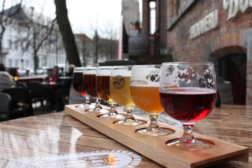 A imagem retrata uma bela apresentação de uma degustação de cervejas artesanais, com diferentes estilos servidos em copos de degustação, dispostos sobre uma base de madeira própria para beer flight (bandeja de amostras).