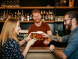 Mulher loira brindando cerveja com homem de óculos e barba, dentro de um bar enquanto o barman observa ao fundo.