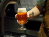 Copo de cerveja artesanal âmbar sendo servido de uma torneira, segurado por uma pessoa com tatuagem no antebraço.