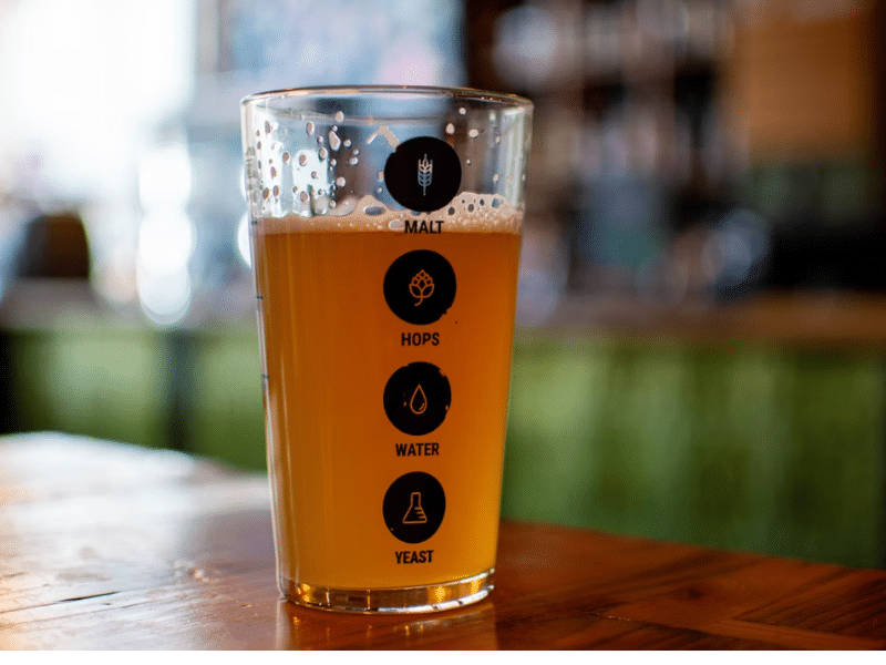 A imagem mostra um copo de cerveja com uma cor dourada, com uma espuma visível na parte superior. O copo possui ícones e rótulos que representam os principais ingredientes da cerveja, organizados de forma vertical.