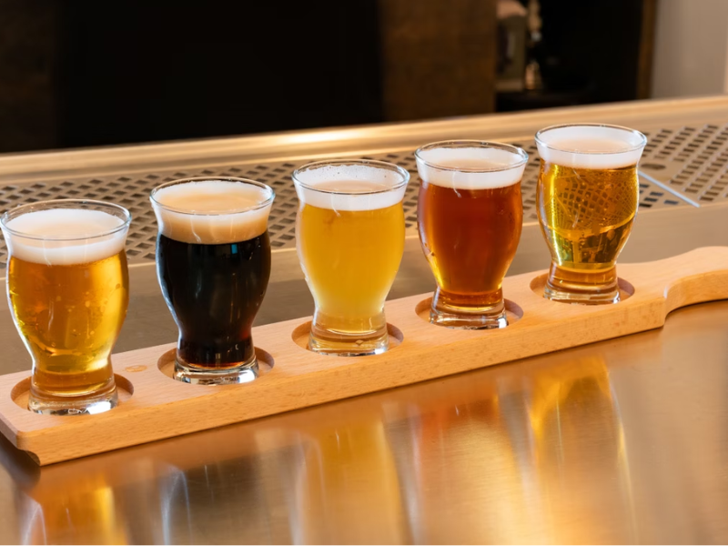 A imagem mostra uma seleção de cinco copos de cerveja na escala SRM organizados em uma tábua de degustação de madeira clara, dispostos sobre uma bancada metálica.