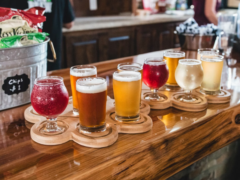 A imagem mostra uma seleção de oito copos de degustação de cervejas na escala SRM organizados em dois suportes de madeira de formato orgânico, colocados sobre um balcão de madeira envernizada em um ambiente de bar ou cervejaria artesanal.