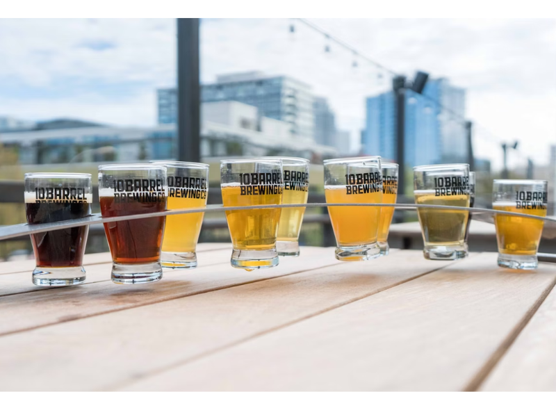 A imagem mostra uma bela sequência de sete copos de degustação de cerveja artesanal, organizados em uma prancha inclinada de madeira, com o logo "10 BARREL BREWING CO." impresso nos copos. O cenário é ao ar livre, provavelmente em um rooftop ou terraço de bar com vista para prédios modernos e céu parcialmente nublado.