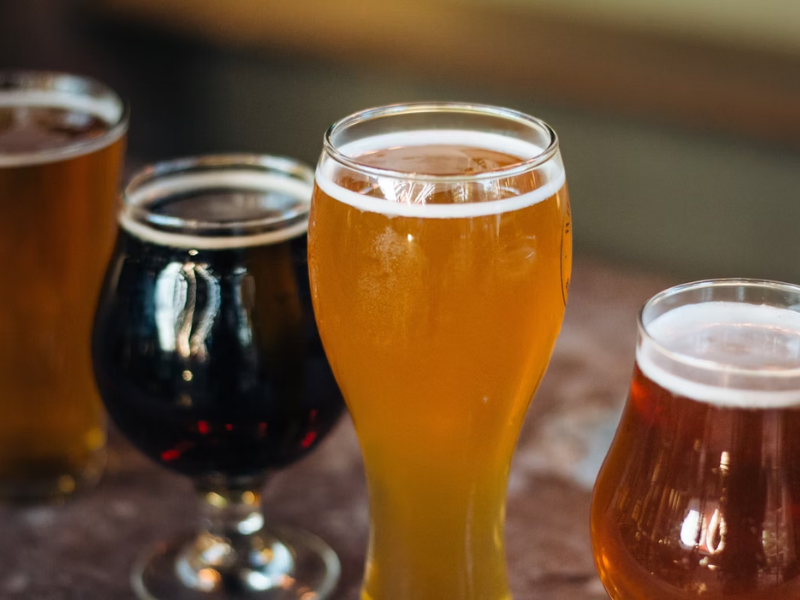 Esta imagem mostra quatro copos de cerveja artesanal, cada um com um estilo e coloração diferente, sugerindo uma variedade de estilos. Eles estão dispostos sobre uma superfície escura, e o foco está no copo central, que é o mais claro. A cena remete a uma experiência de degustação ou a um ambiente de pub/bar artesanal.