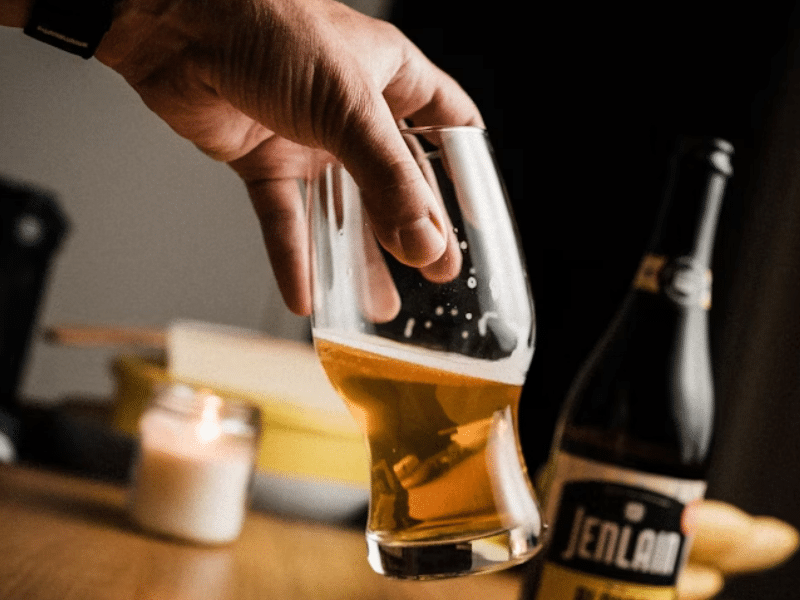 A imagem mostra uma mão segurando um copo de cerveja parcialmente cheio, em um ambiente interno com iluminação suave e aconchegante. A cerveja tem uma coloração dourada clara e parece estar levemente gaseificada. O copo tem um formato curvado, elegante, ideal para degustação de cervejas artesanais.