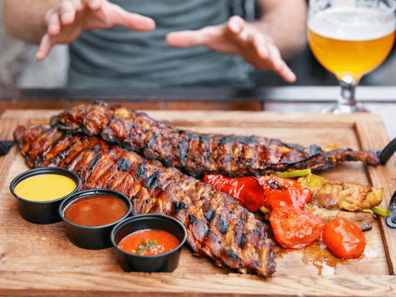 A imagem mostra uma mão segurando um copo de cerveja parcialmente cheio, visto em destaque no primeiro plano. A cerveja tem uma coloração dourada, com uma leve espuma esbranquiçada nas bordas do líquido. O copo é de vidro, com design moderno e curvado, ideal para degustação, e está sendo inclinado como se estivesse prestes a ser levado à boca.