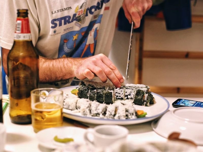 Claro, com base na imagem que você enviou, aqui está uma descrição:
A foto mostra um close-up focado nas mãos de uma pessoa que está cortando rolos de sushi em uma tábua ou prato branco. Os rolos de sushi, que parecem ser do tipo uramaki (arroz por fora), estão dispostos em um prato.
Na lateral esquerda da cena, há uma garrafa de cerveja de vidro âmbar quase cheia e um copo de cerveja com um líquido amarelo claro e espumante, sugerindo que a pessoa está a desfrutar de uma bebida enquanto prepara ou come a refeição.