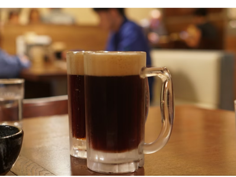 A imagem mostra duas canecas de vidro com uma bebida escura e espumosa, possivelmente uma cerveja, sendo servidas em uma mesa de restaurante ou bar. As canecas têm uma espuma densa e cremosa no topo, criando uma divisão clara entre o líquido escuro e a espuma.