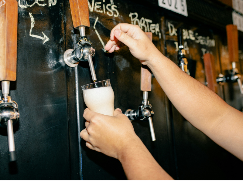 A imagem mostra uma pessoa servindo cerveja diretamente de uma torneira de chope em um ambiente de bar ou microcervejaria. A pessoa segura um copo de vidro, que está sendo preenchido com uma espuma densa e cremosa.