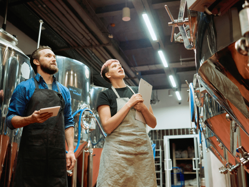 Dois cervejeiros, um homem com barba e tablet e uma mulher com cabelo rosa e prancheta, inspecionando tanques de fermentação de aço inoxidável em uma cervejaria industrial.