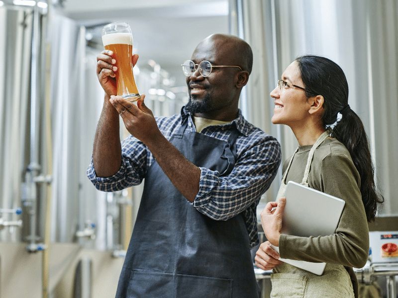Dois cervejeiros, um homem e uma mulher, em aventais, sorrindo enquanto inspecionam a cor e a clareza de uma cerveja em um copo alto em frente a tanques de fermentação de aço inoxidável.