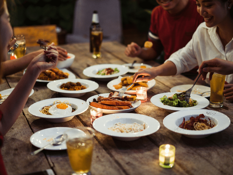 Alt Text (Sucinto):
Grupo de pessoas reunidas em torno de uma mesa rústica de madeira, compartilhando diversas porções de comida e bebidas em um jantar informal com luz de velas.
