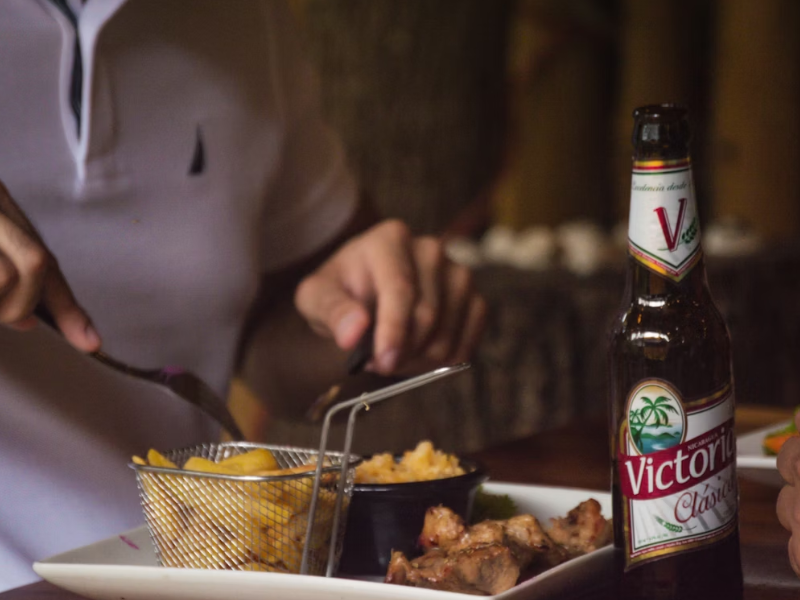 A imagem em questão apresenta uma cena de refeição em um ambiente fechado e com iluminação baixa, focada em uma garrafa de cerveja e um prato de comida, com uma pessoa ao fundo.