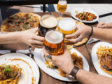 A imagem captura uma cena vibrante e sociável de harmonização de cervejas e comidas em uma mesa ao ar livre, provavelmente em um ambiente de cervejaria artesanal ou restaurante descontraído.