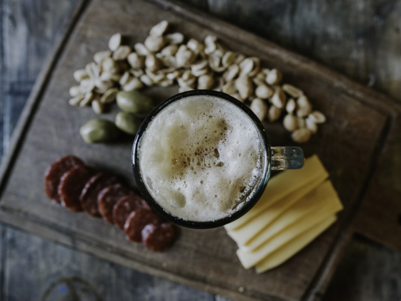 A imagem mostra uma tábua rústica de madeira com uma caneca de cerveja clara e espuma cremosa no centro.