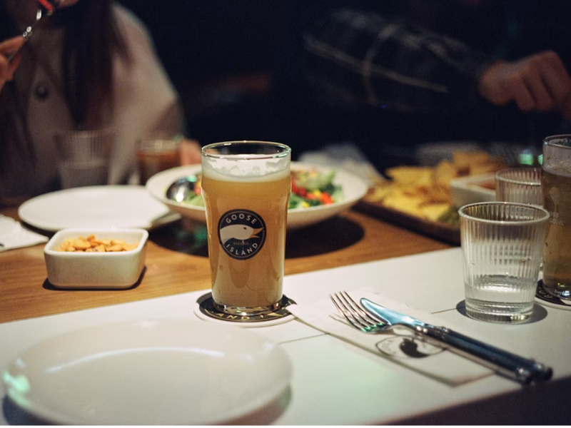 A imagem mostra uma mesa posta em um ambiente de bar ou restaurante, com um copo de cerveja artesanal clara no centro.