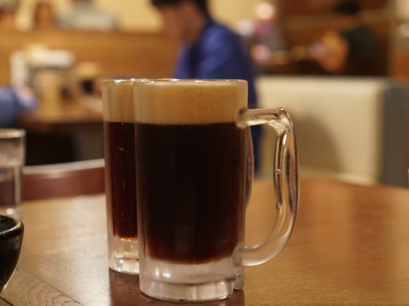 A imagem mostra duas canecas de cerveja escura por conta de um dos tipos de malte, servidas sobre uma mesa de madeira em um ambiente que parece ser um restaurante ou bar. As canecas têm alças largas e espessas, com paredes de vidro grosso, típicas para servir cervejas mais encorpadas.