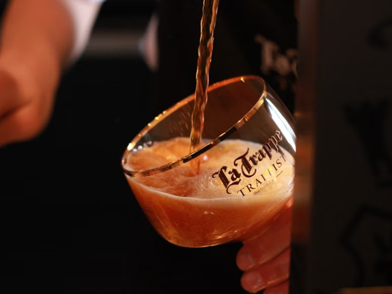 A imagem mostra uma taça sendo enchida com uma cerveja âmbar ou dourada escura de um dos tipos de malte, que está sendo servida diretamente de uma torneira (provavelmente em um bar ou evento cervejeiro). A taça está inclinada para facilitar o serviço e reduzir a formação excessiva de espuma.